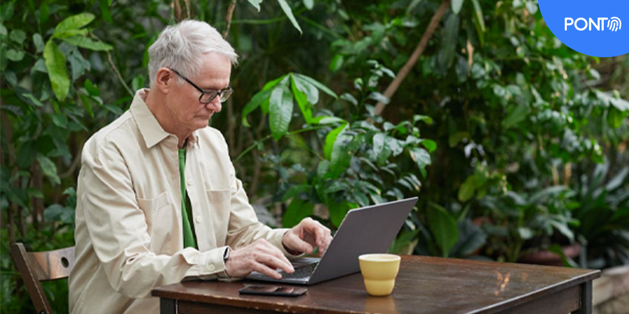 Homem idoso sentado em uma mesa ao ar livre, mexendo em um computador.