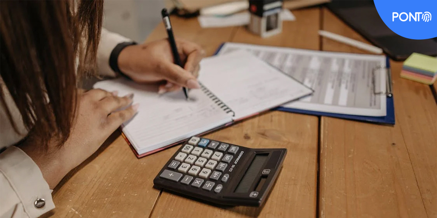 Imagem de uma mulher escrevendo em um caderno com uma calculadora em um lado e uma prancheta no outro.